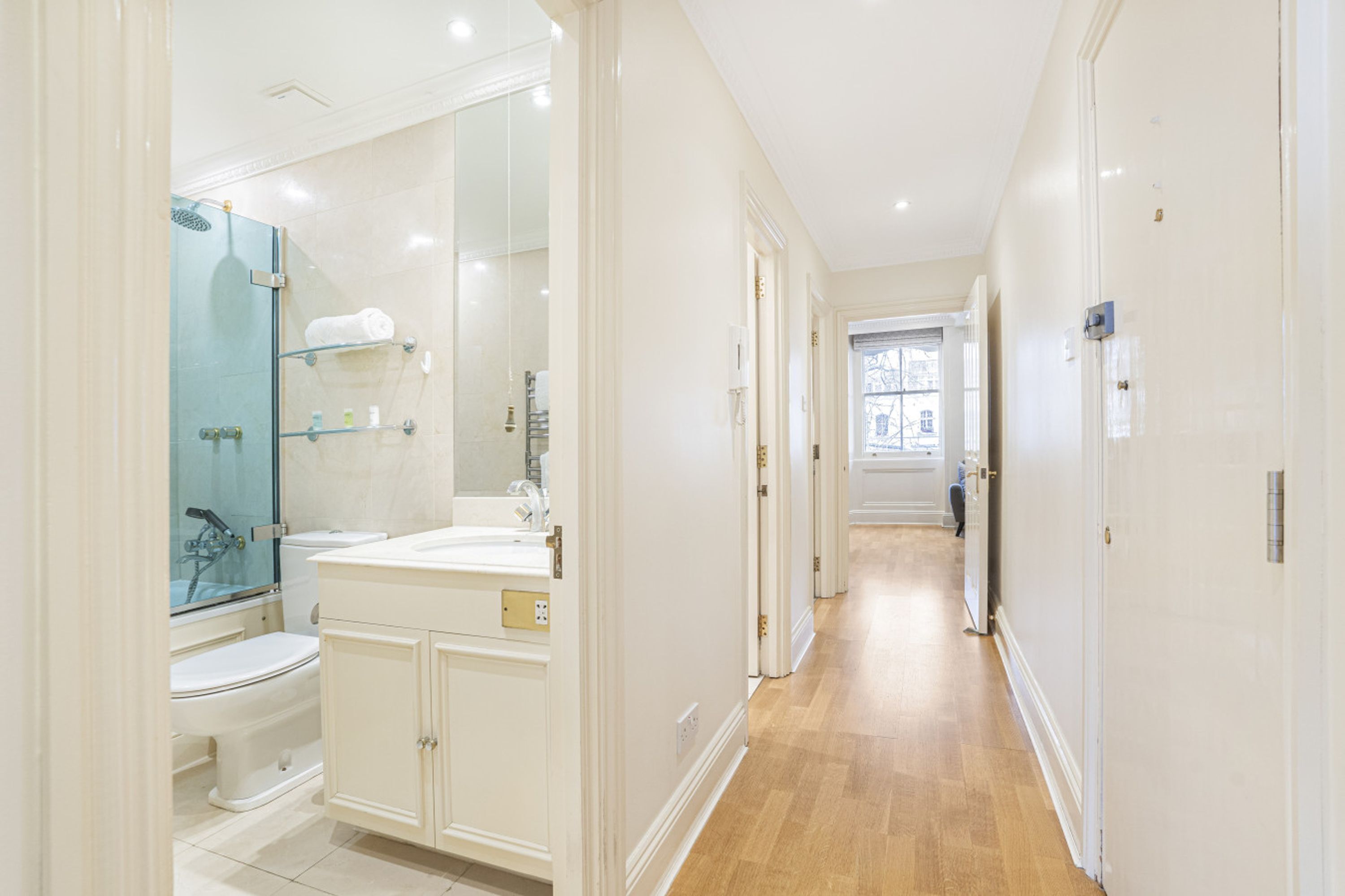 A modern bathroom with a glass shower enclosure, a white sink, and a toilet. A hallway leads to another room.