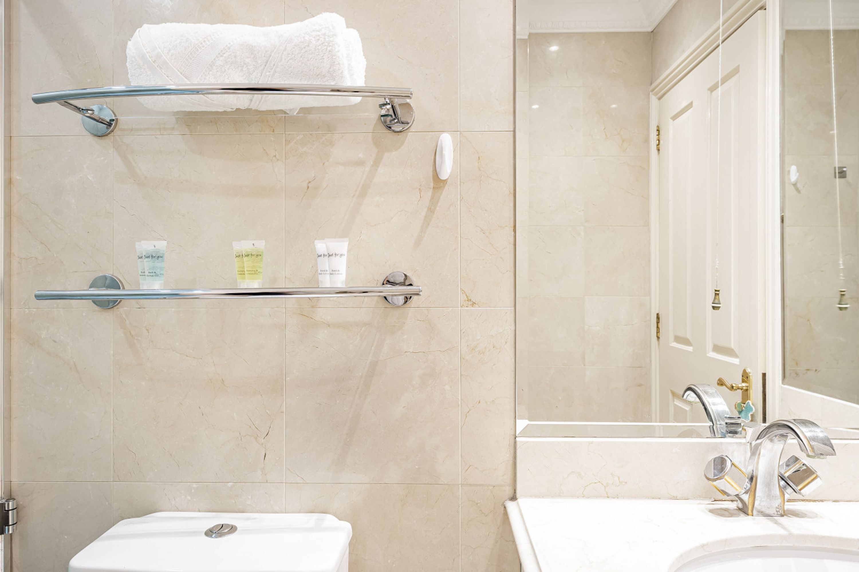 A bathroom with a toilet, a sink, and a mirror. There are two towel racks and a shelf with toiletries.