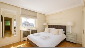 A hotel bedroom with a large bed, two bedside lamps, a view of the city through a window, and a walk-in closet. - Luxury Knightsbridge Home Next To Hyde Park (London)