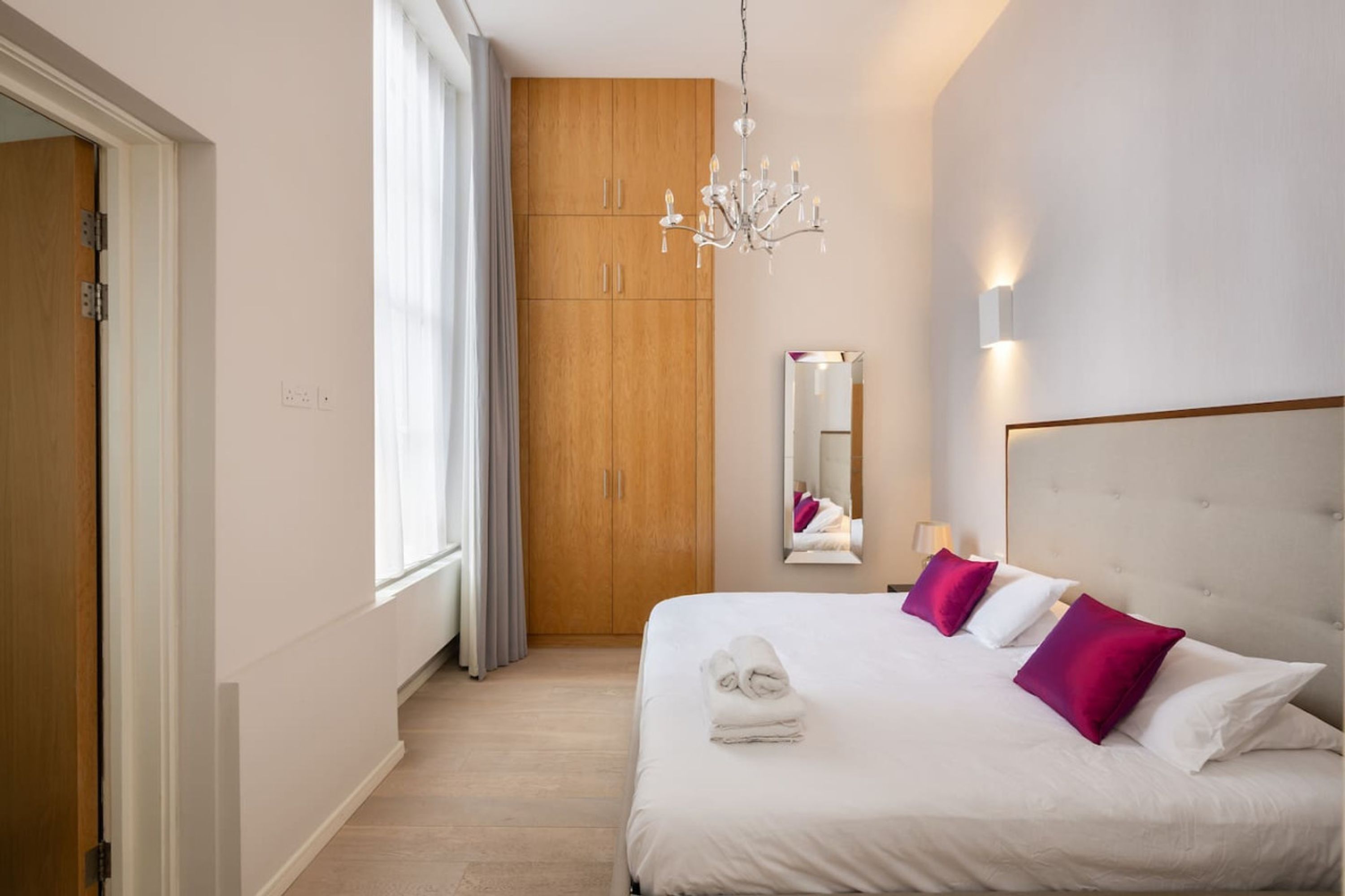 Habitación de hotel con una cama grande, un armario de madera, una araña, un espejo y una ventana con cortinas.