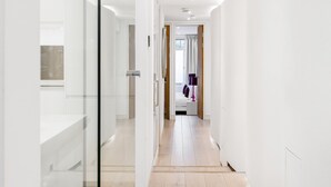 Corredor moderno e bem iluminado, com piso de madeira e divisórias de vidro, que leva a um quarto com cama e janela.