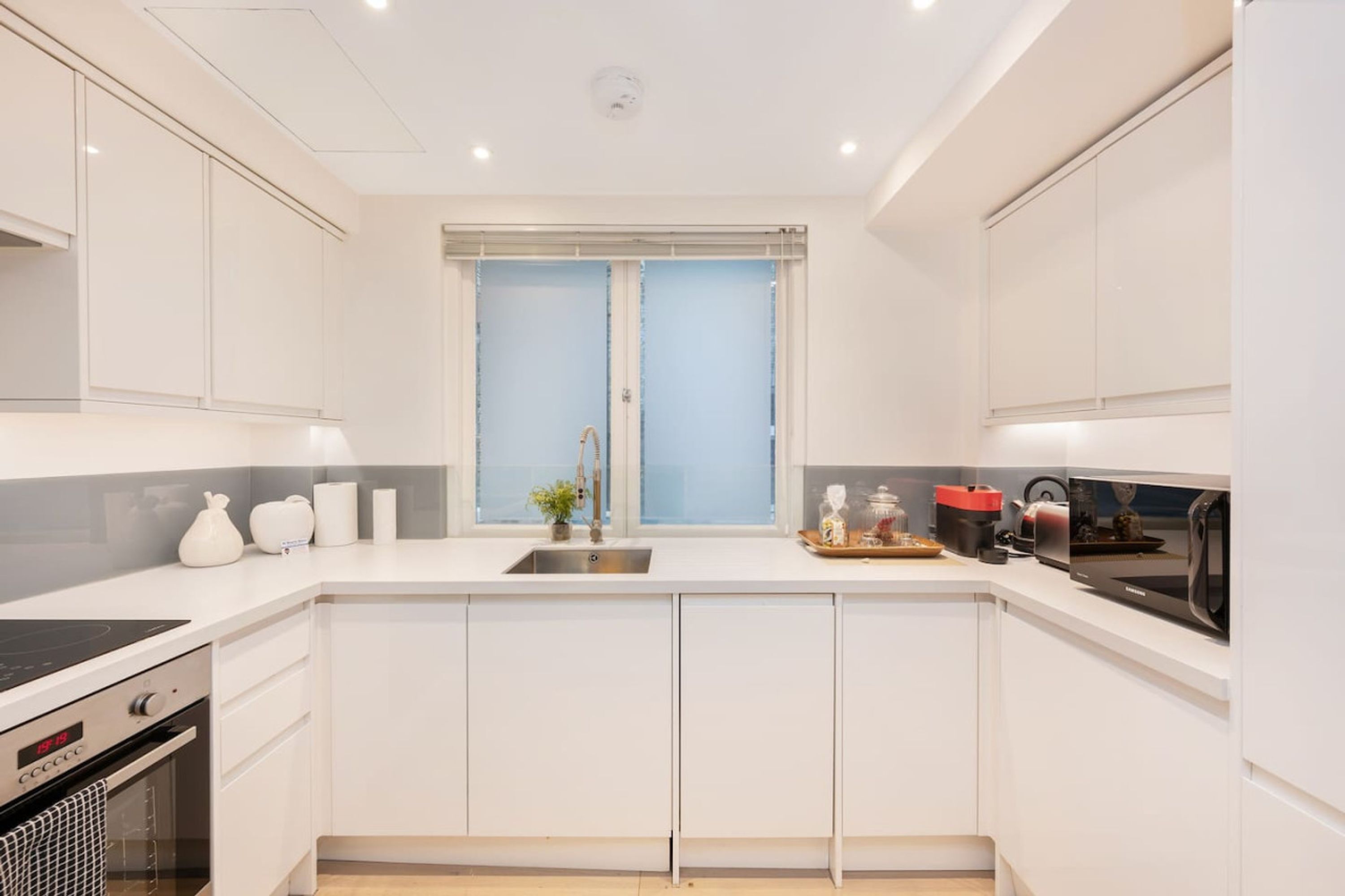 A modern kitchen with white cabinetry, a built-in oven, a sink, and a window with blinds.