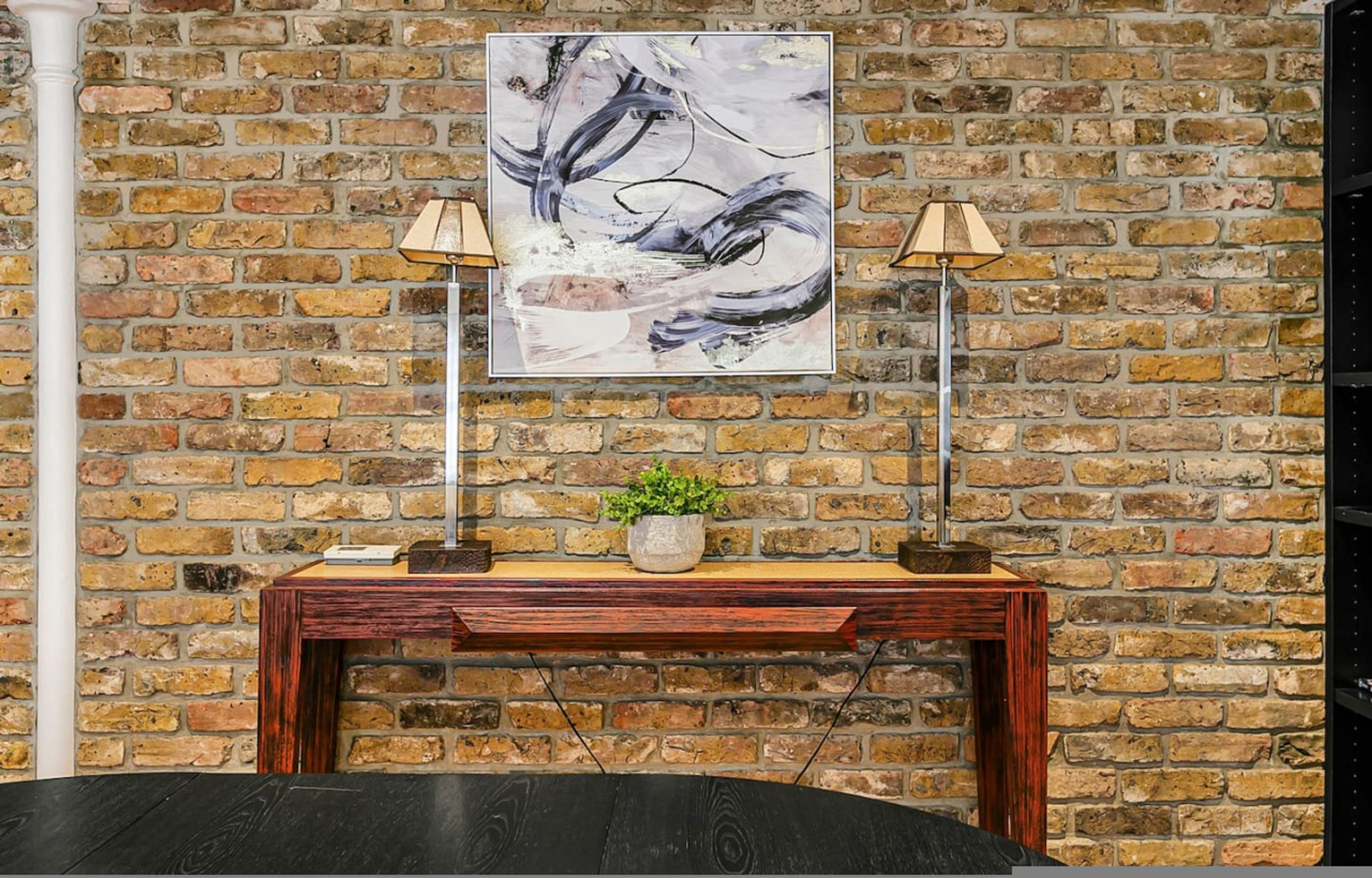 A brick wall with a framed abstract painting, a wooden console table with a potted plant, and two floor lamps.