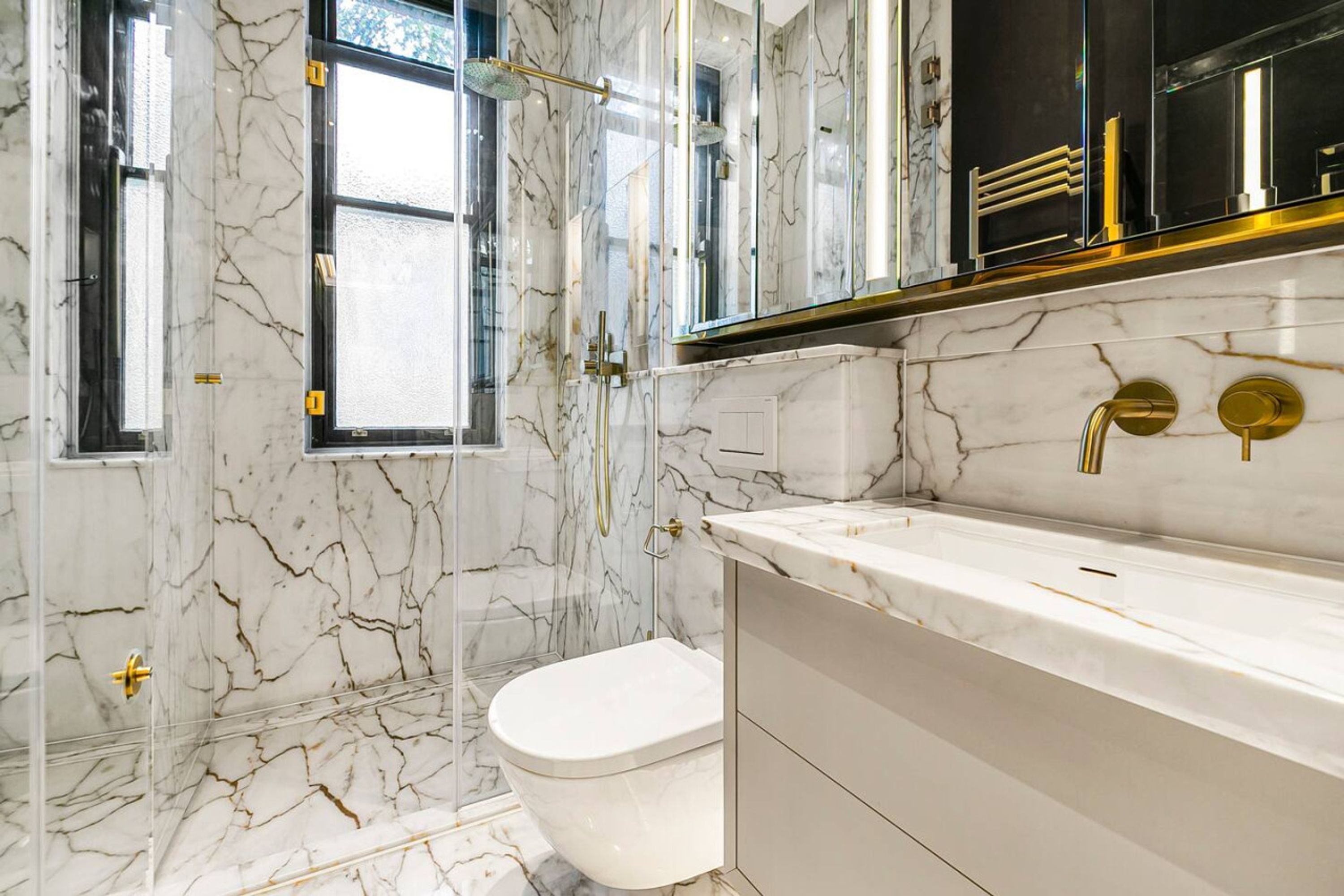 A bathroom with marble walls, a glass shower enclosure, a white toilet, and a gold-finished sink.
