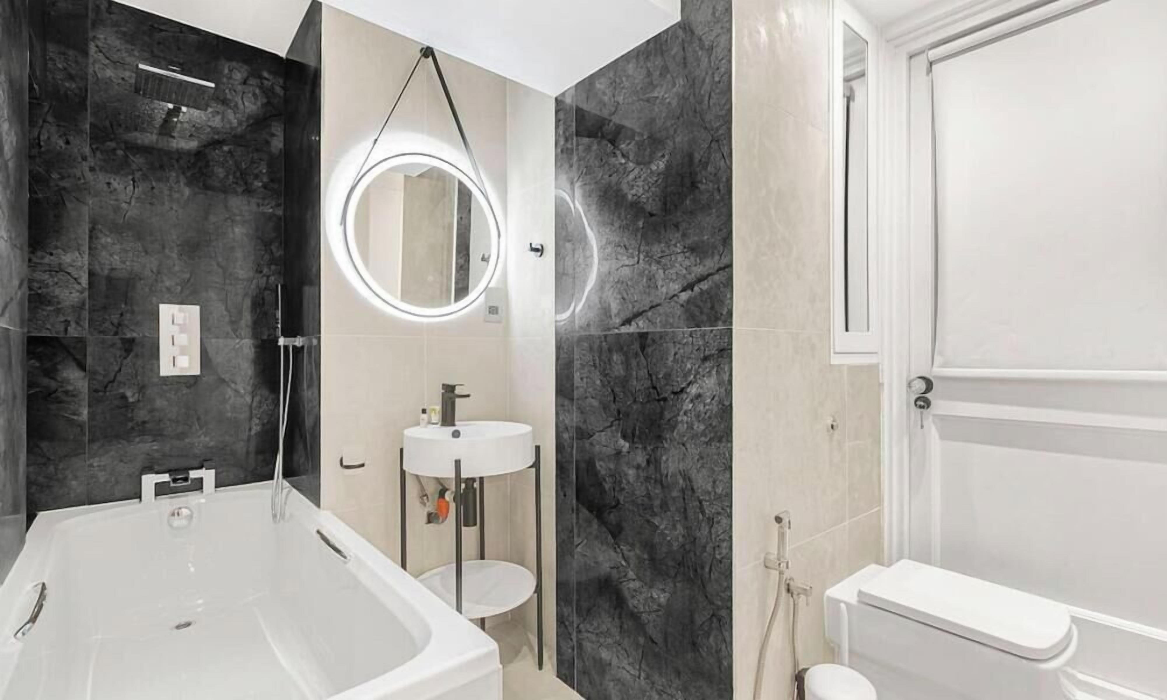 A modern bathroom with a freestanding bathtub, a round mirror with a light, a pedestal sink, and a toilet.