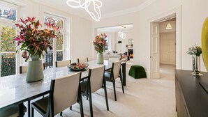 Ein moderner Essbereich mit einem großen Tisch, Stühlen und zwei Blumenvasen. Ein Balkon mit Geländer und eine Tür zu einem anderen Raum sind zu sehen.