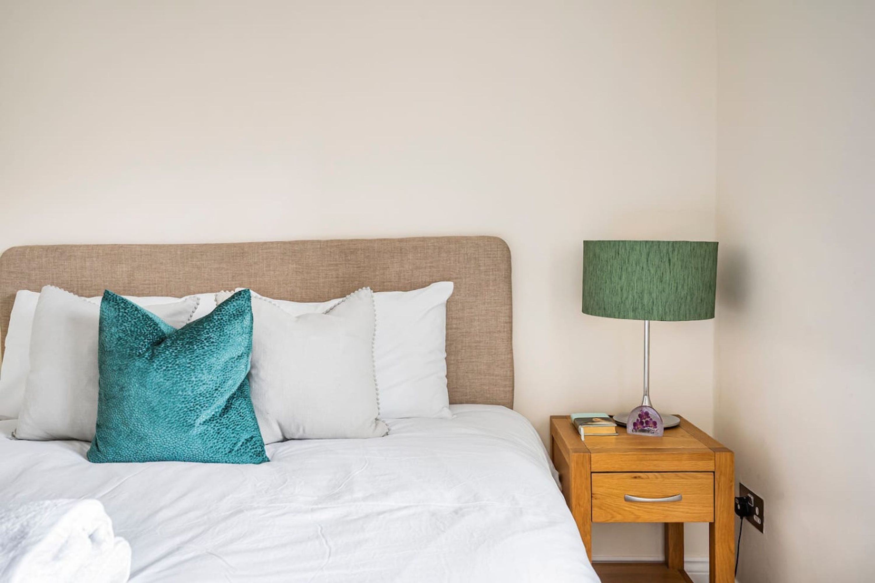 Un lit bien fait, avec du linge de lit blanc et un coussin turquoise, une table de chevet en bois avec une lampe verte et un mur sobre.