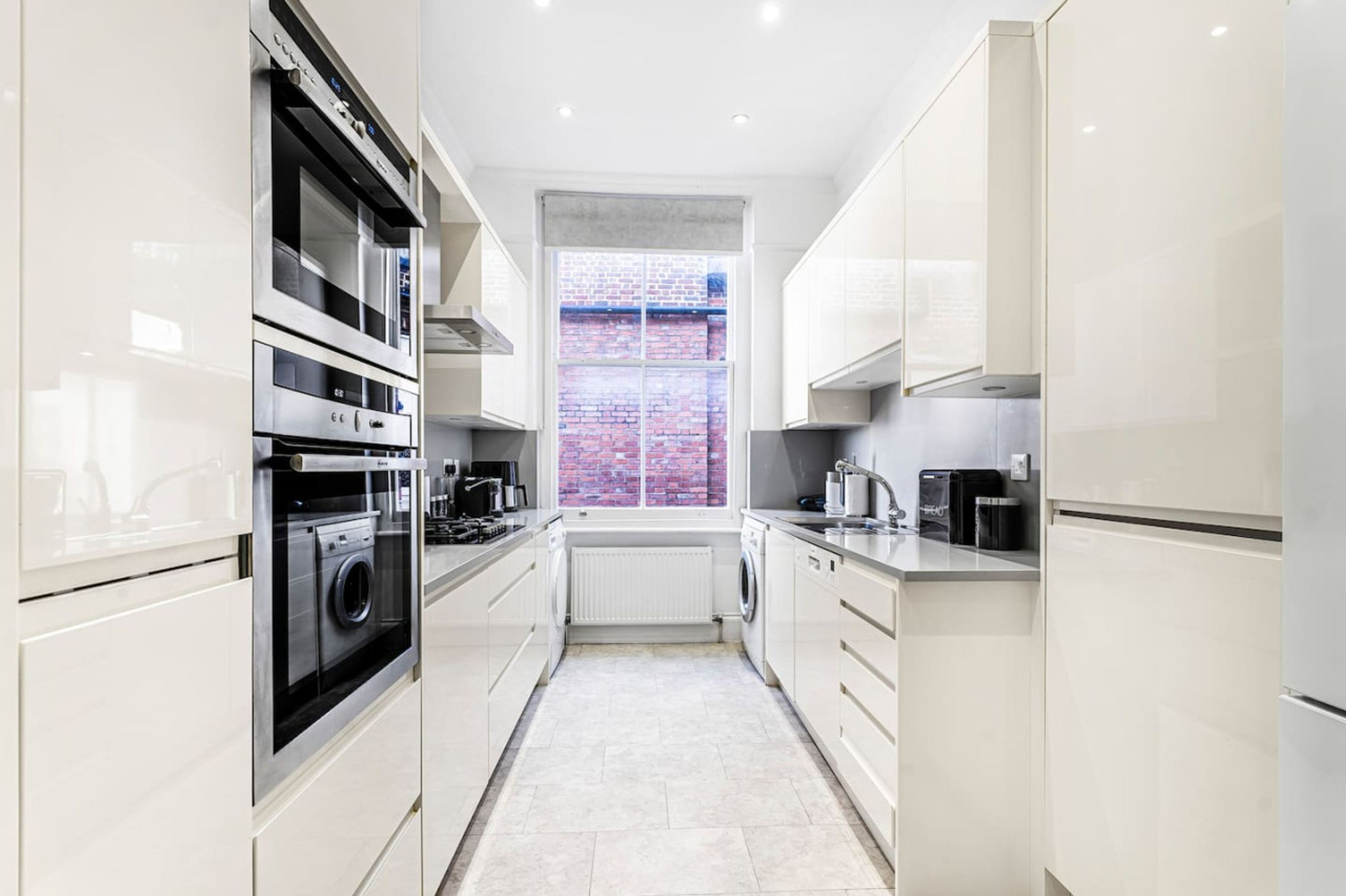 Een moderne keuken met roestvrijstalen apparatuur, een raam met uitzicht op een bakstenen muur en witte kasten.