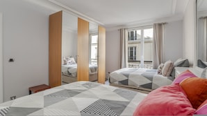 A modern bedroom with two beds, a large mirror, and a window with a view of buildings. - Luxury 4 Bed Residence - Next to Arc de Triomphe (Paris)