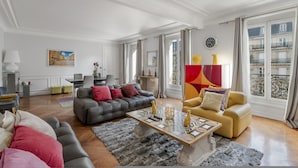 A spacious living room with a grey sofa, a yellow armchair, and a coffee table with decorative items. There is a painting on the wall and a window with curtains. - Luxury 4 Bed Residence - Next to Arc de Triomphe (Paris)