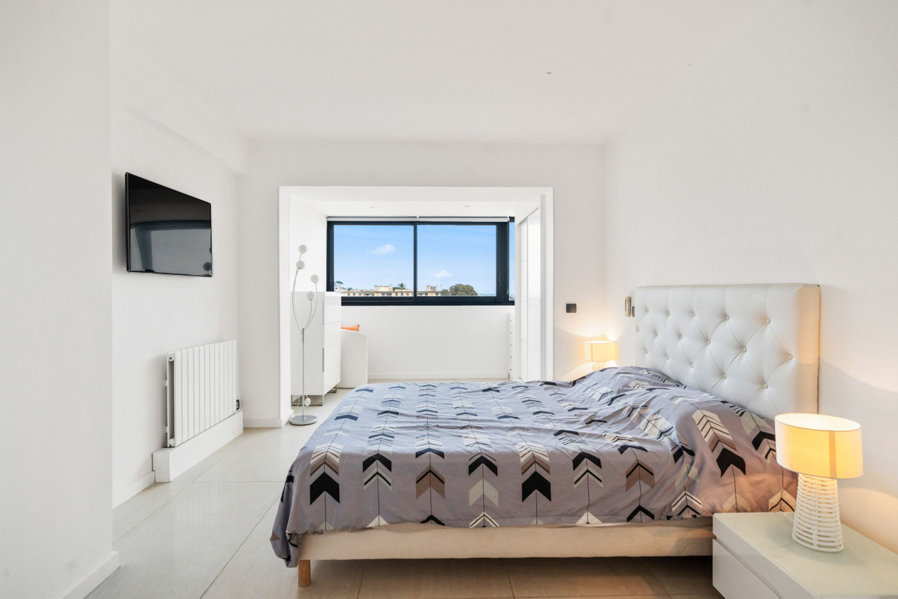 A modern bedroom with a large bed, a window with a city view, a flat-screen TV mounted on the wall, and a bedside table with a lamp.