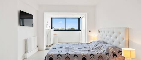 A modern bedroom with a large bed, a window with a city view, a flat-screen TV mounted on the wall, and a bedside table with a lamp.