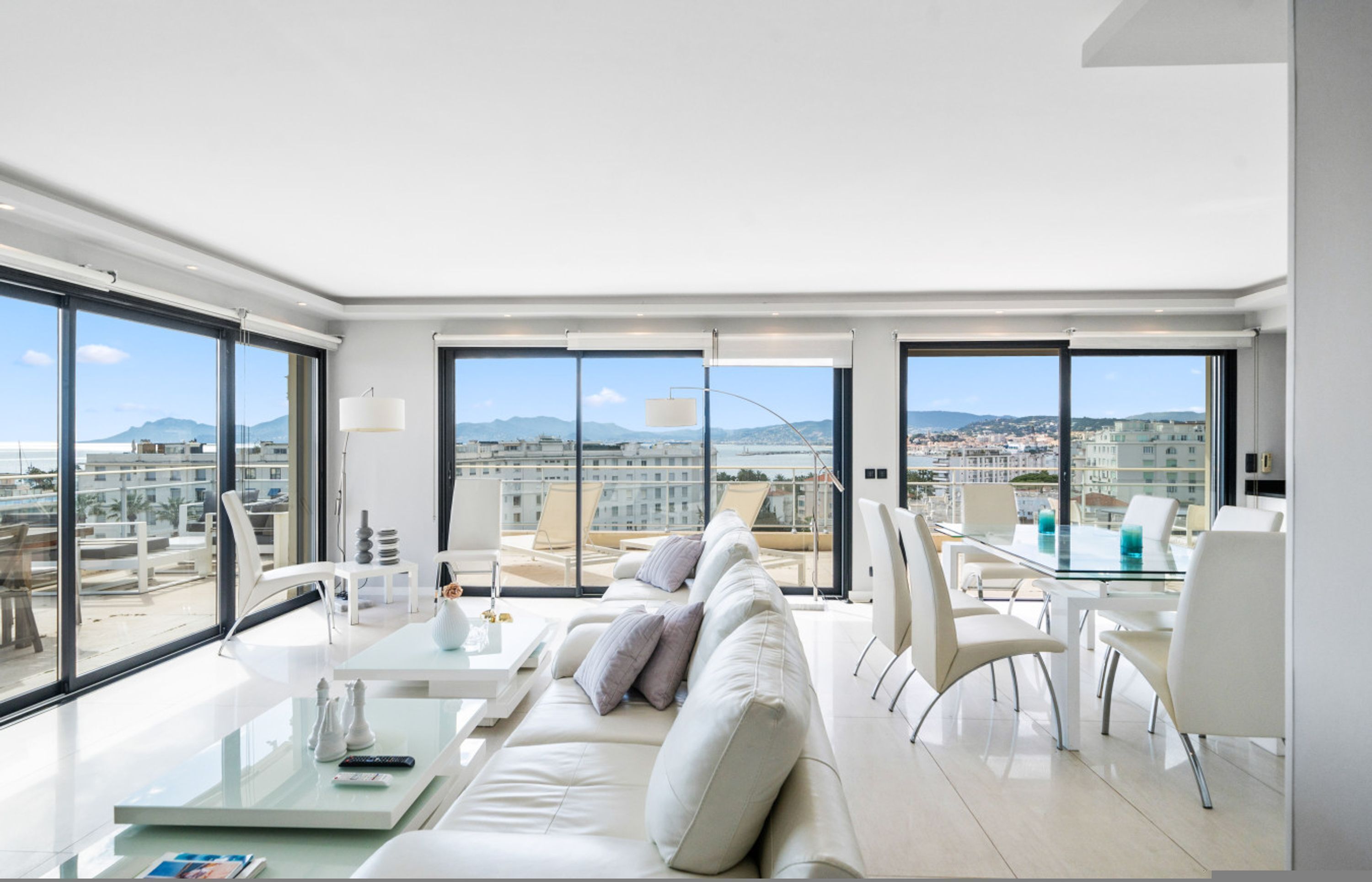 A modern living room with a glass table, white sofa, and large windows offering a city view.