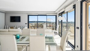 Zona pranzo moderna con tavolo in vetro e sedie bianche, televisore a schermo piatto sulla parete e vista sulla città attraverso ampie finestre.