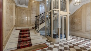 A grand staircase with ornate railings and patterned carpet leading to a glass elevator. - Luxury 5 Bed Residence - Opposite Arc de Triomphe (Paris)