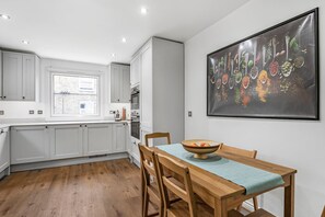 A modern kitchen with white cabinets, a wooden dining table, and a large window. - Luxury Modern 5-bed Home Maida Vale (London)