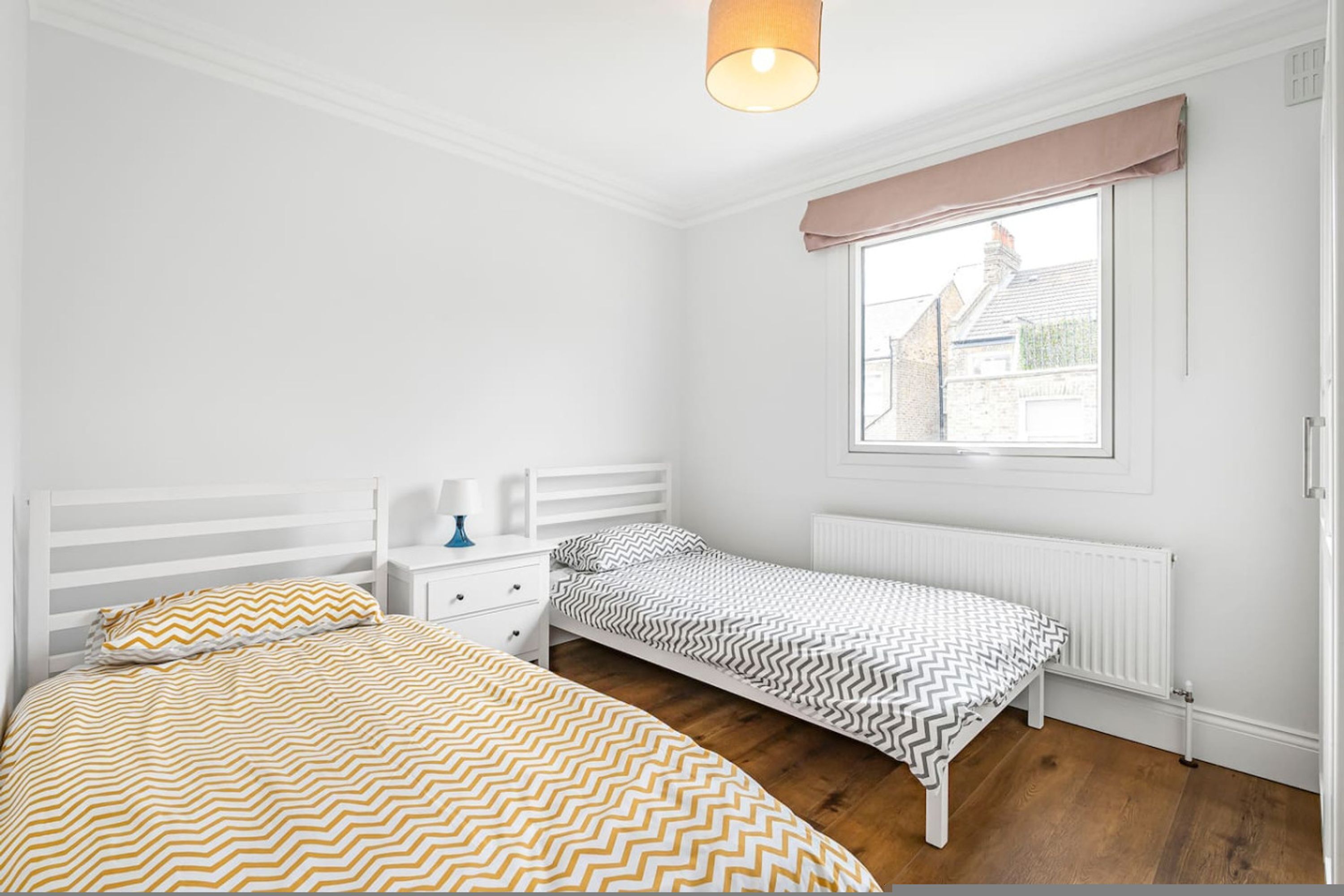 A bedroom with two single beds, a white radiator, a window with a view of buildings, and a white ceiling.