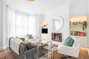 A modern living room with a grey sofa, white armchair, and a coffee table. There is a fireplace with a framed picture above it, and a bookshelf with books and decorative items. - Luxury Modern 5-bed Home Maida Vale (London)
