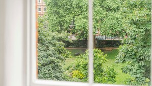 A view through a window of a lush garden with a pond, surrounded by dense foliage and a building in the background. - Stylish 1BR Kensington Home With Balcony (London)