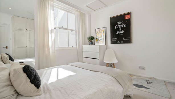A bedroom with a large bed, a bedside table with a lamp, a window with curtains, a wall-mounted poster, and a door leading to another room. - 2-bed Knightsbridge Home (London)