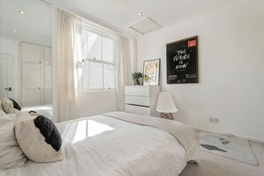 Une chambre avec un grand lit, une table de chevet avec une lampe, une fenêtre avec des rideaux, une affiche accrochée au mur et une porte donnant sur une autre pièce.