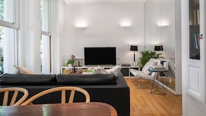 A modern living room with a black sofa, wooden chairs, a dining table, a flat-screen TV, and a bookshelf. - 2-bed Knightsbridge Home (London)