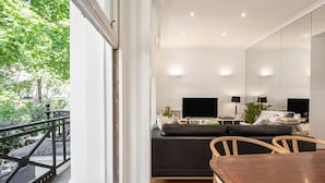 A modern living room with a large window, a black sofa, a wooden dining table, and a kitchen area with two black monitors. - 2-bed Knightsbridge Home (London)