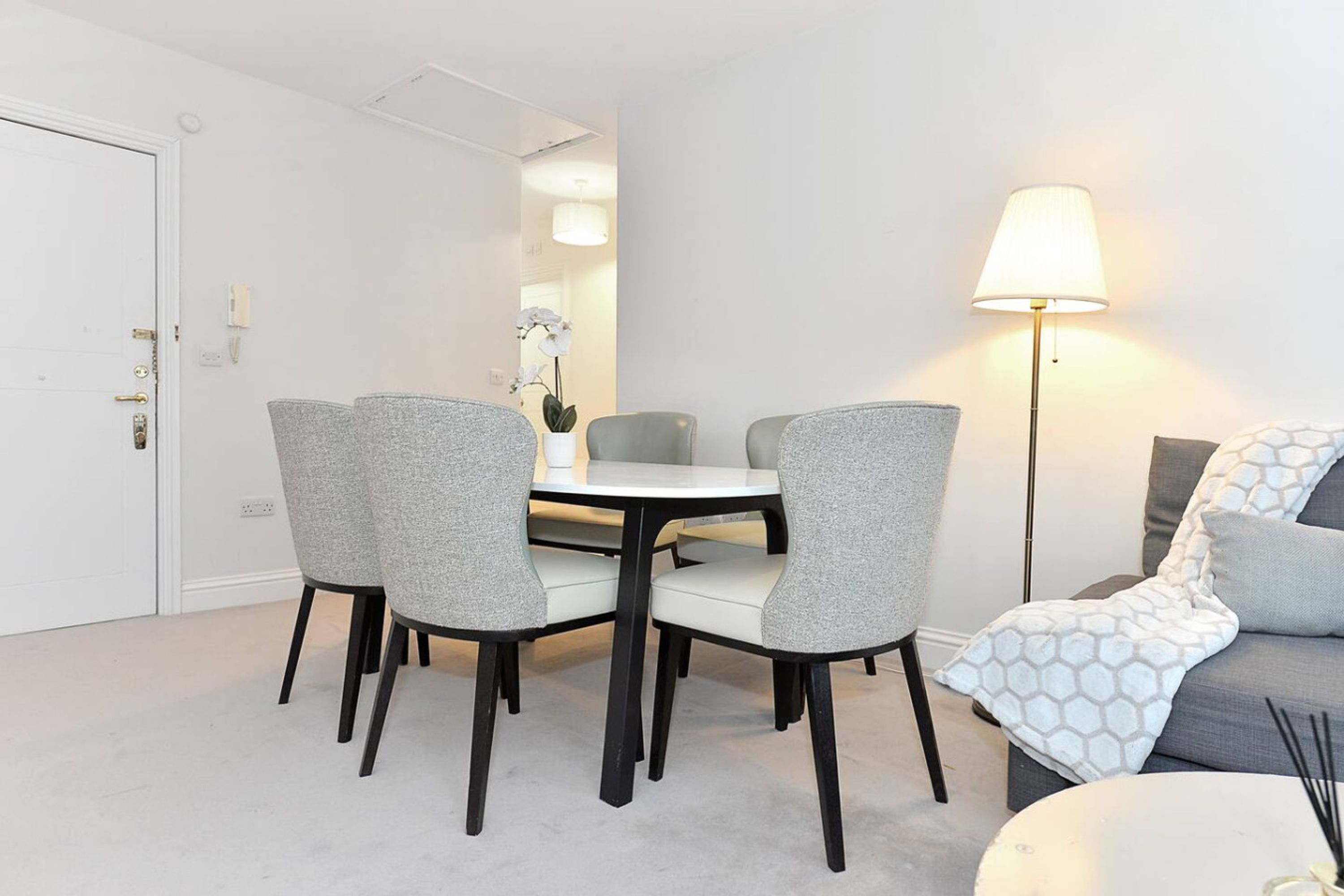 A dining area with a round table and four grey chairs, a sofa with patterned cushions, and a floor lamp.
