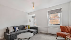 A modern living room with a grey sofa, a round coffee table, and a red armchair. There is a window with a view of buildings outside. - Cadogan Square Penthouse (London)