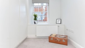 A small, white-walled room with a radiator, a window, and a suitcase on the floor. - Cadogan Square Penthouse (London)