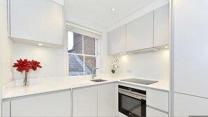 A modern kitchen with white cabinetry, a built-in oven, and a window overlooking a brick building. - Cadogan Square Penthouse (London)