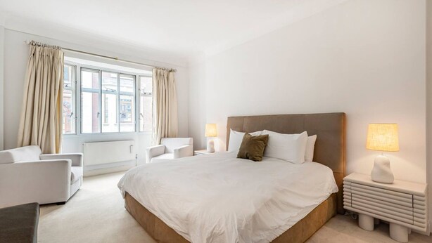 A modern hotel room with a large bed, two bedside tables with lamps, a sofa, and a view of the city through large windows. - Luxury 3BR Mayfair Apartment Near Green Park (London)