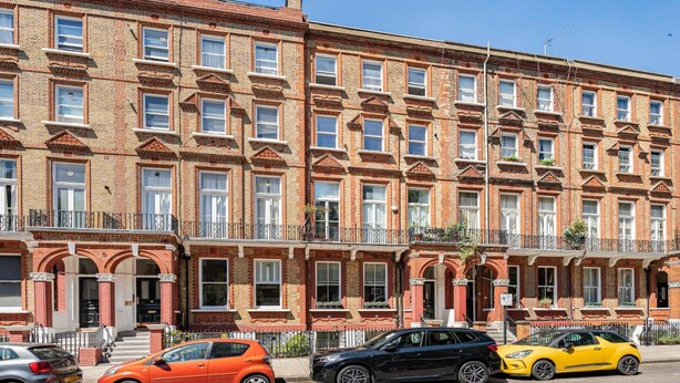 Une rangée de maisons de ville aux façades en briques rouges, avec des fenêtres et des balcons identiques, des voitures garées dans la rue et un ciel bleu dégagé.