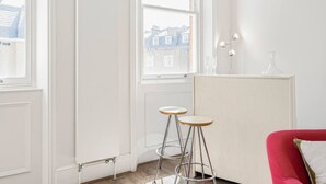 Une pièce lumineuse avec une chaise rouge, deux tabourets, un radiateur et une fenêtre donnant sur des immeubles.