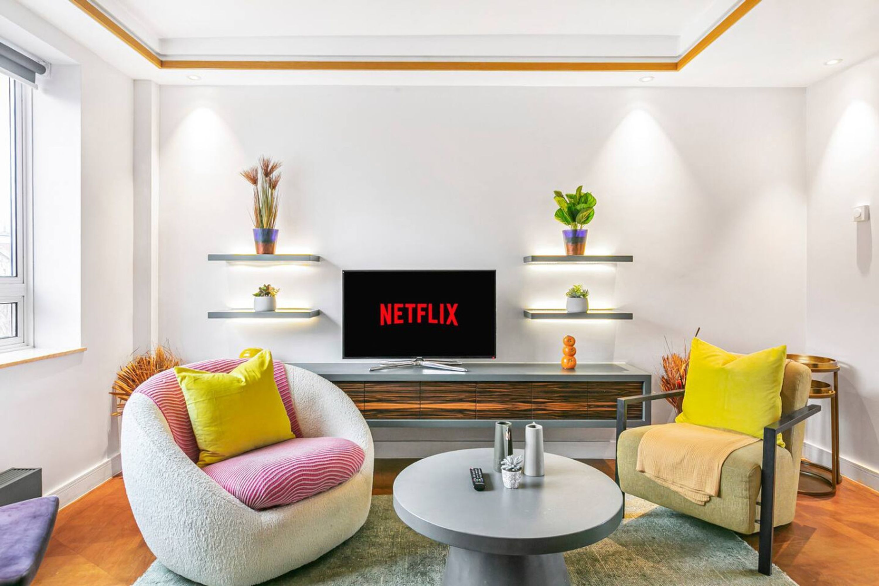 A modern living room with a flat-screen TV, two armchairs, a coffee table, and decorative plants on shelves.