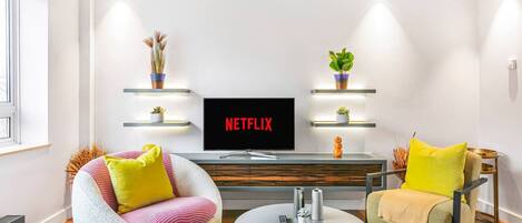A modern living room with a flat-screen TV, two armchairs, a coffee table, and decorative plants on shelves.