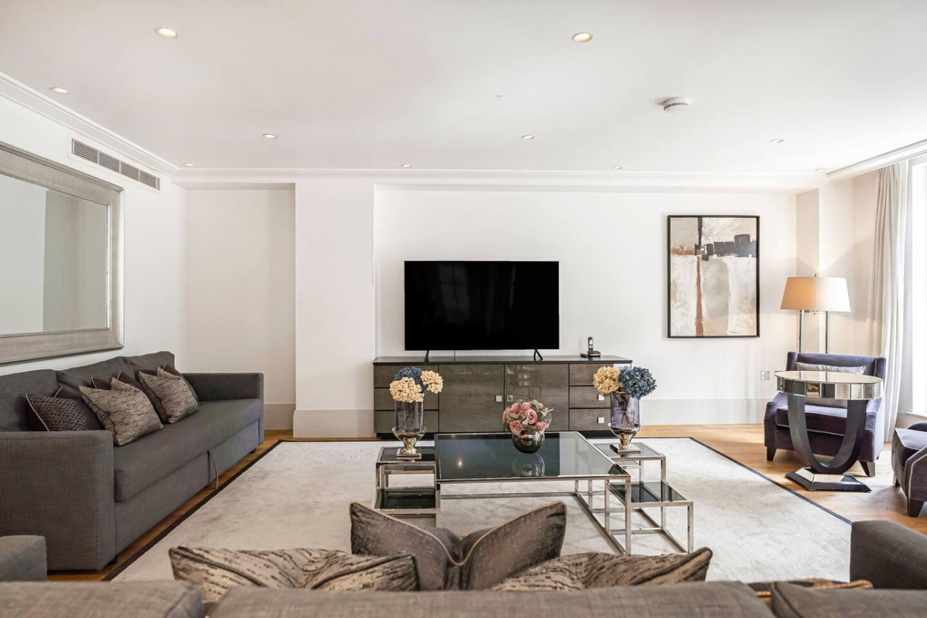A modern living room with a grey sofa, a glass coffee table, and a flat-screen TV mounted on the wall.