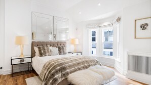 Un dormitorio con una cama grande, dos mesitas de noche, un espejo, un ventanal con vistas y un radiador.