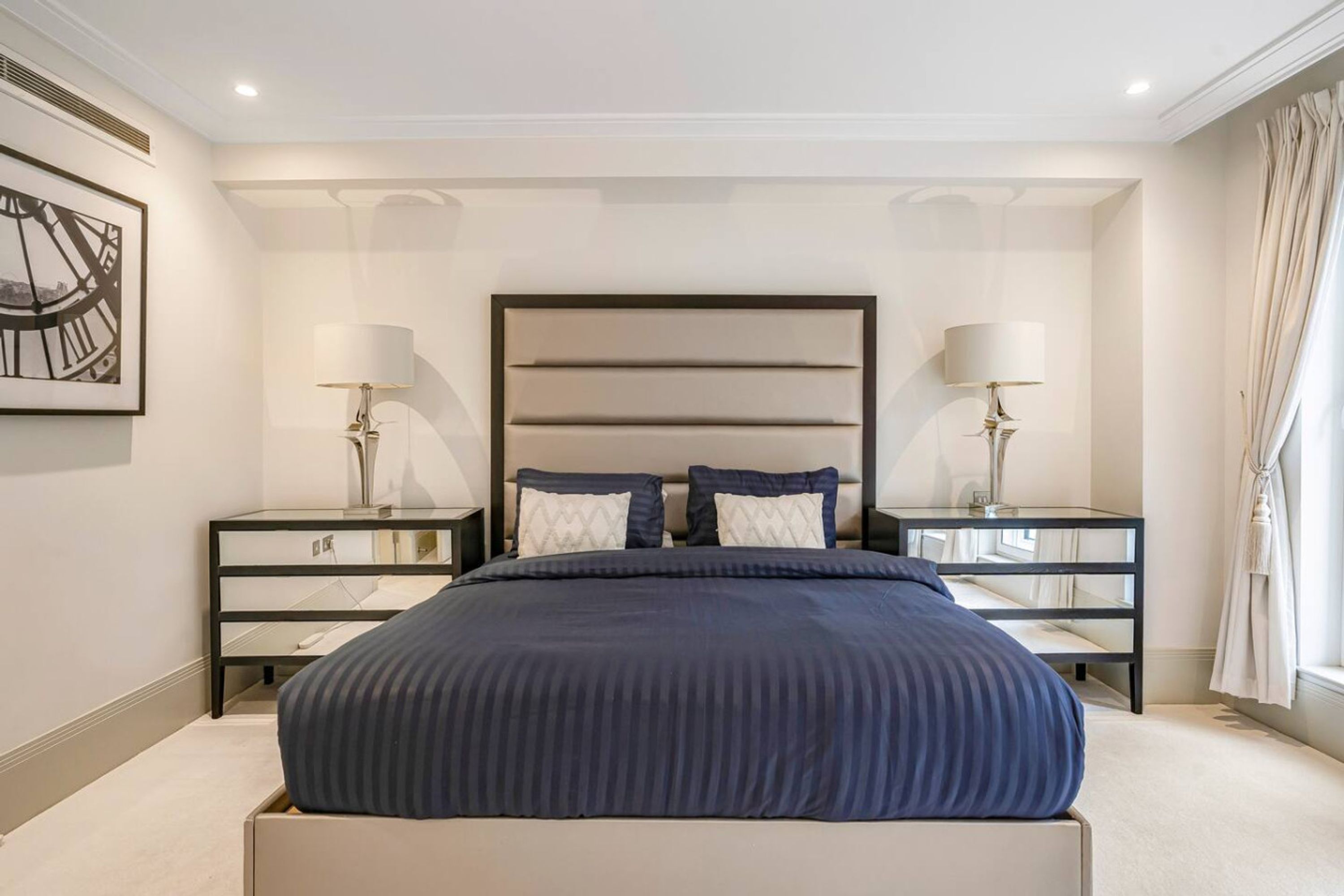 Um quarto moderno com uma cama grande, duas mesinhas de cabeceira e uma janela ampla com cortinas.