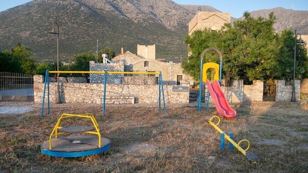 Ein Spielplatz mit Rutsche und Schaukeln, vor einer Kulisse aus Steinbauten und Bergen.