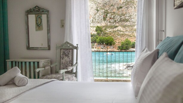 A bedroom with a bed, a chair, a small table, and a view of the sea through a balcony. - Gerolimin Blue (East Mani)