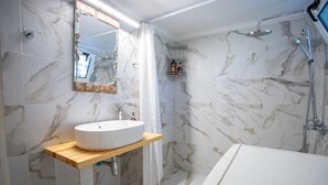A bathroom with a marble-tiled shower, a white sink on a wooden shelf, and a mirror with a decorative frame. - Gerolimin Blue (East Mani)