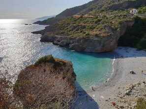 Un paisaje costero con playa rocosa, aguas turquesas cristalinas y un pequeño edificio en una colina.