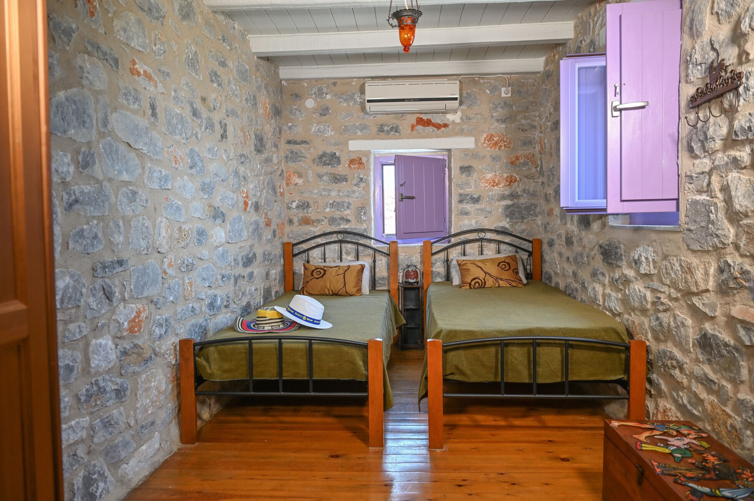 A narrow room with two beds, stone walls, wooden flooring, a purple door, and an air conditioning unit.