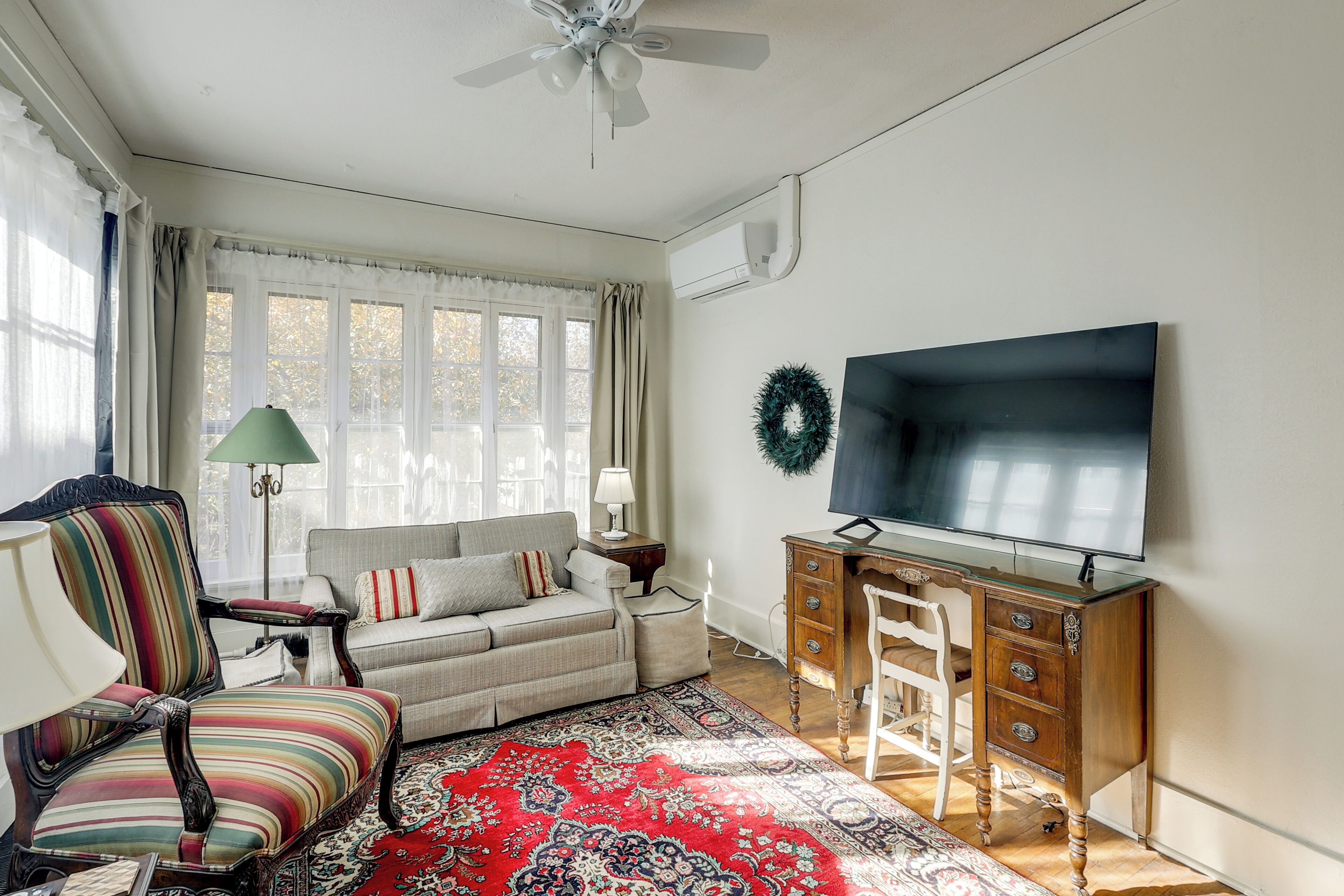 A living room with a sofa, a striped armchair, a wooden desk with drawers, a television, and a large window with curtains.