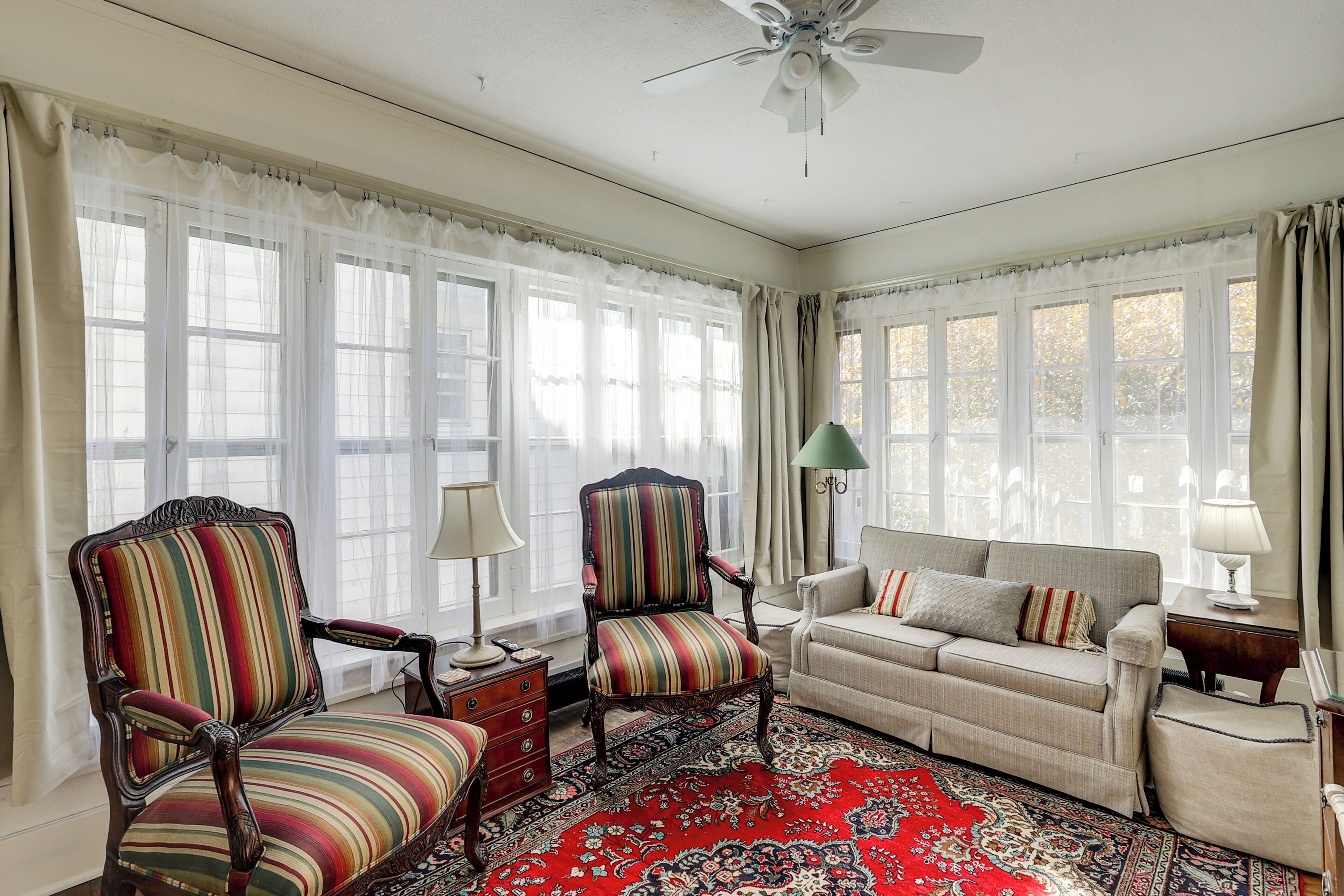 A room with a large window, a ceiling fan, a sofa, two armchairs, a small table, and a patterned rug.
