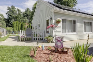 Uma casa com fachada branca, varanda frontal com um banco e um pequeno jardim com cobertura de palha e plantas.