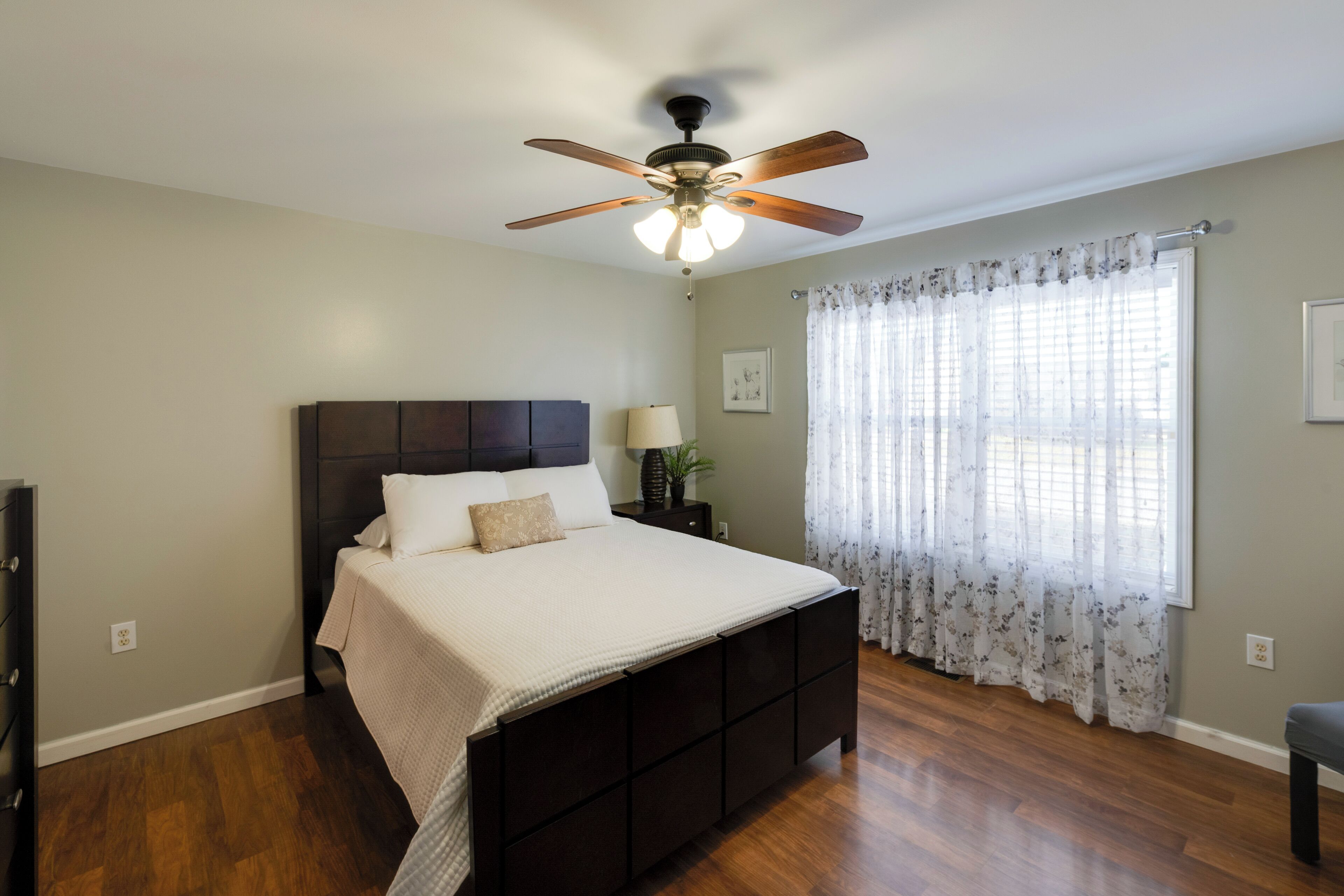 A bedroom with a dark wooden bed, a ceiling fan, a window with floral curtains, and a nightstand with a lamp.