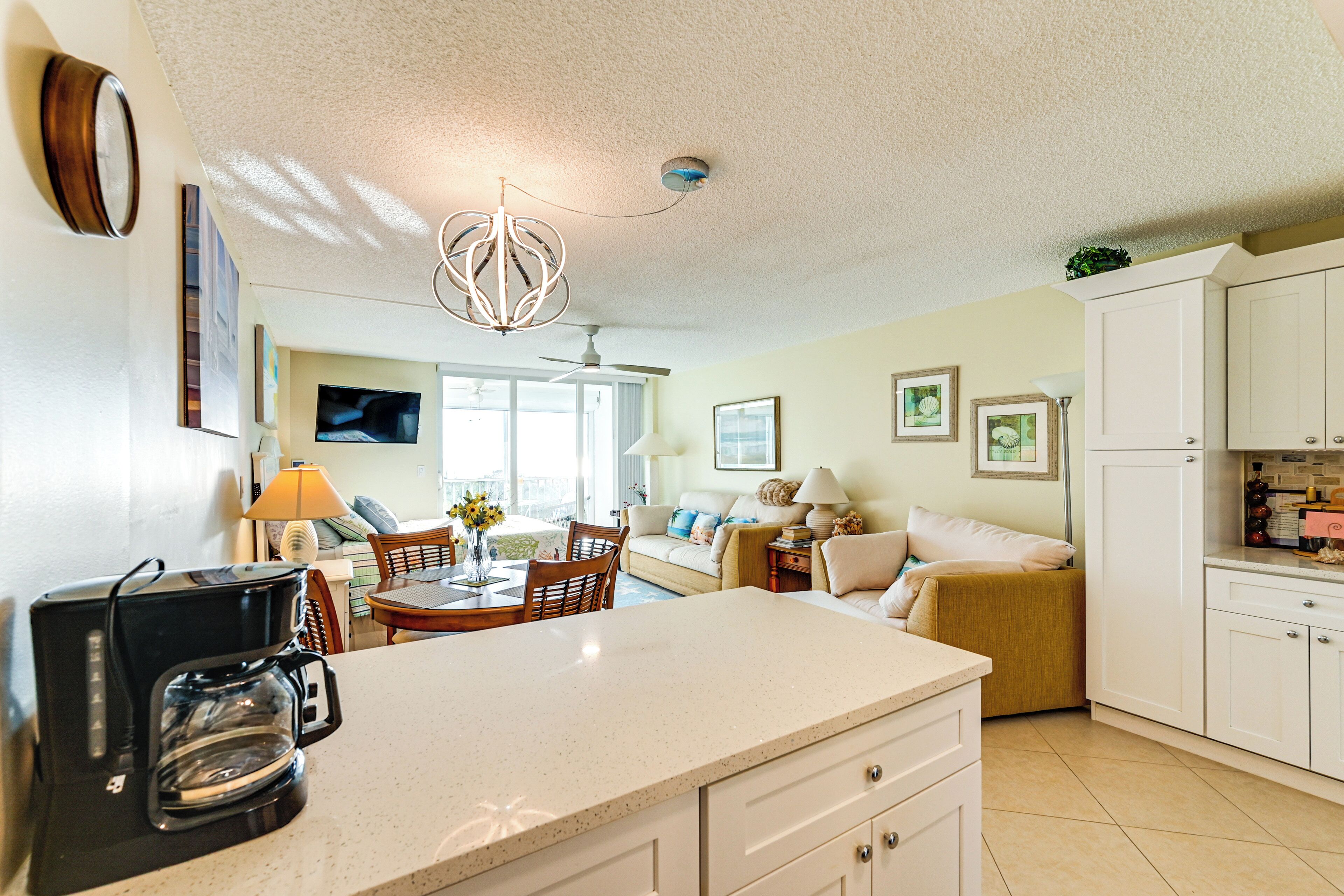 A well-lit kitchen with a coffee maker, a dining area with a table and chairs, and a living room with a sofa and a TV.