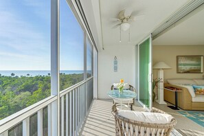 Een balkon met uitzicht op de oceaan en groen, een plafondventilator, een glazen tafel met stoelen en een bank met een schilderij.